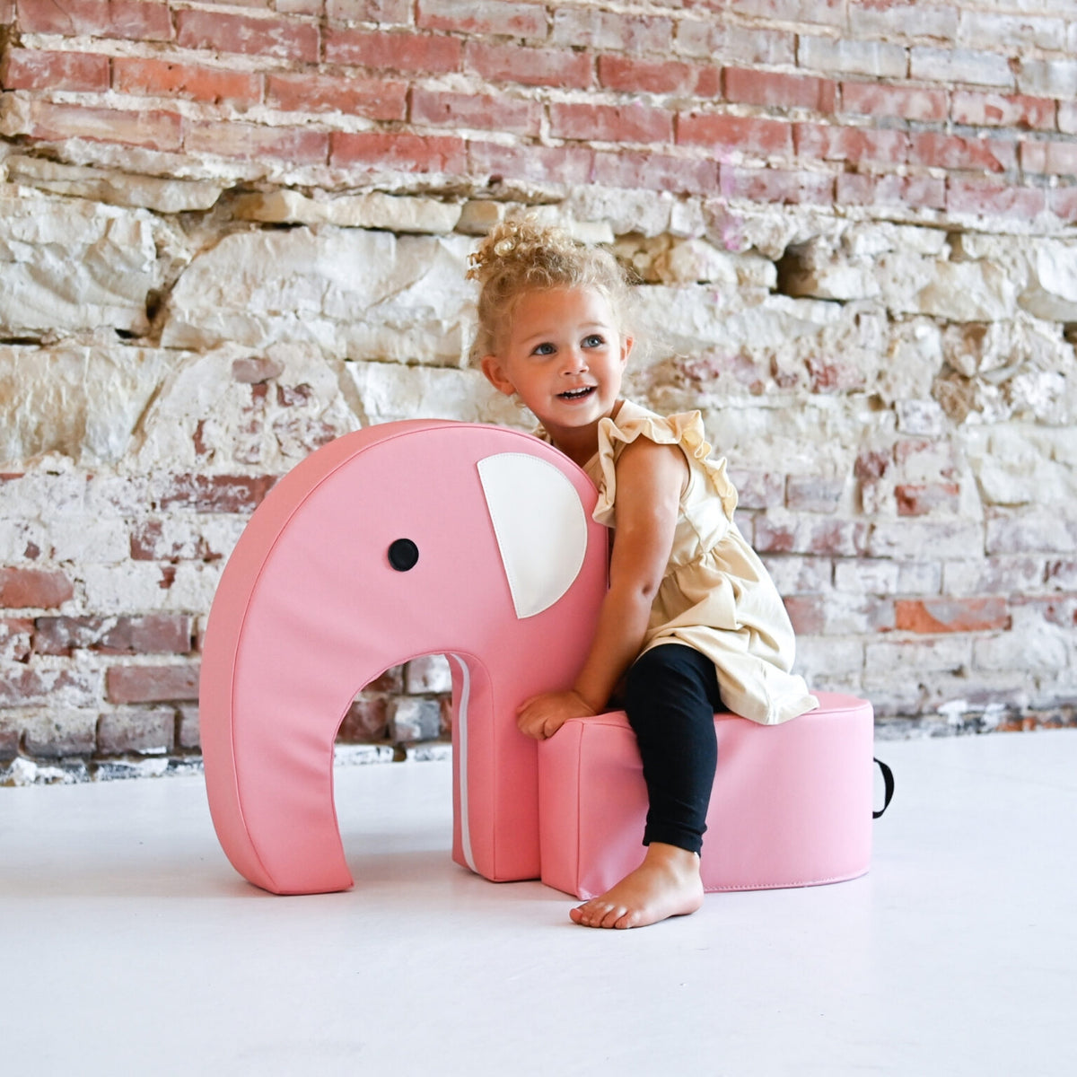 Child sitting on a pink elephant-shaped chair against a brick wall.