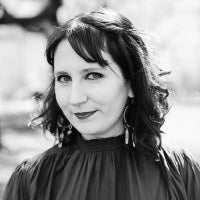 A black and white portrait of a woman with dark hair, wearing a dark high-neck top and dangling earrings, set against a softly blurred outdoor background.
