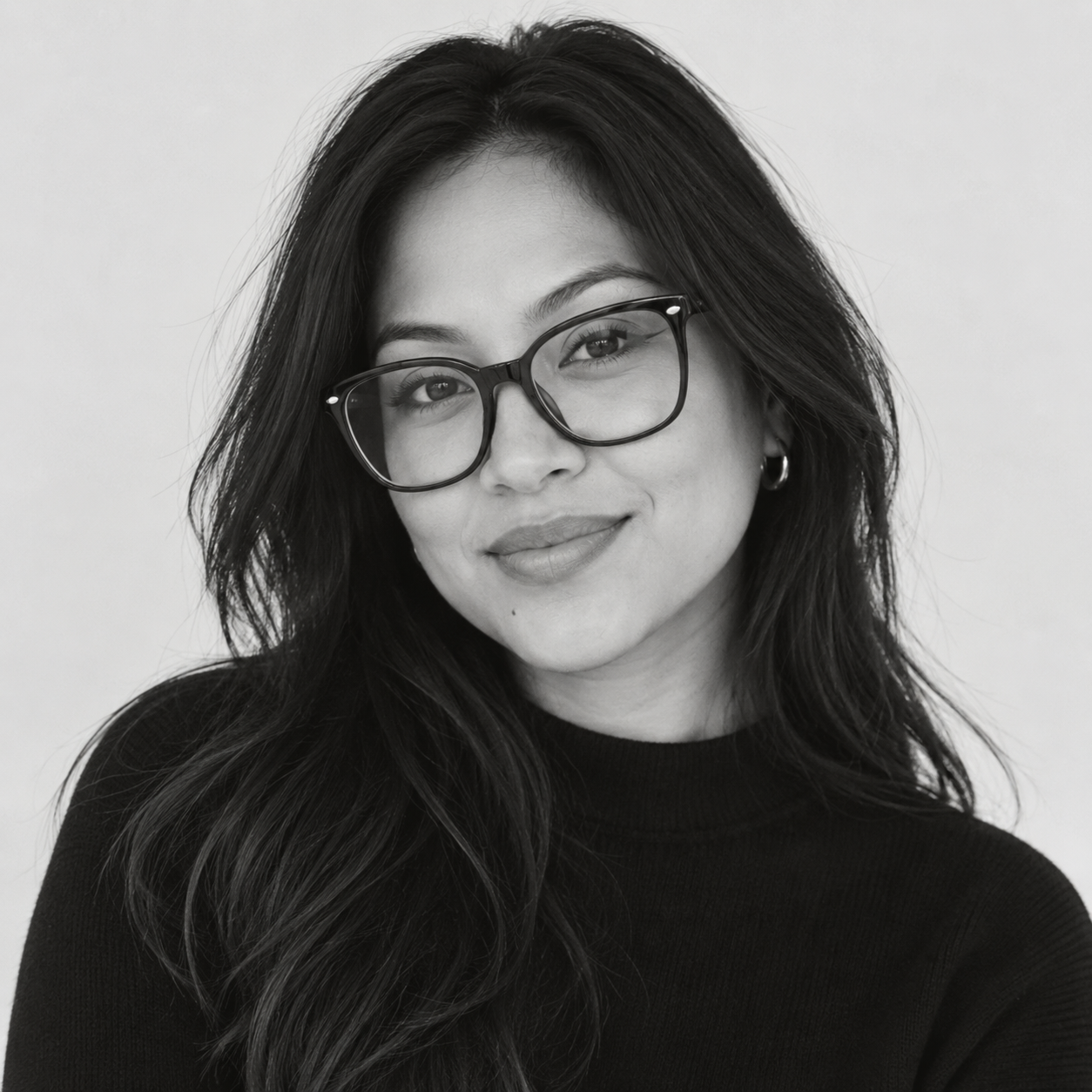 A black and white portrait of a woman with long dark hair wearing thick black rectangular eyeglasses and a black ribbed turtleneck sweater against a light gray background.