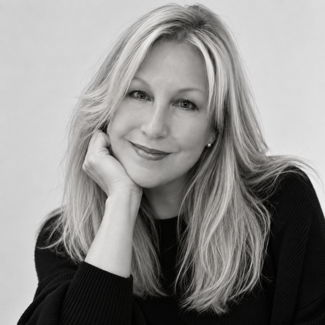 A black and white portrait of a smiling blonde woman resting her chin on her hand, wearing a dark ribbed top against a plain light background.